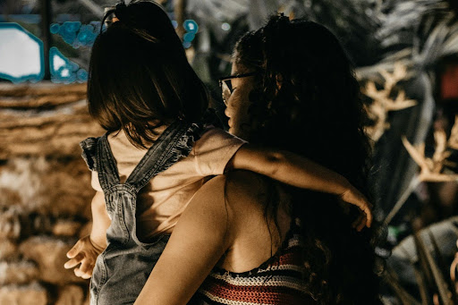 A woman with long curly hair and glasses holds a young child wearing denim overalls, both facing away from the camera in a warmly lit setting.