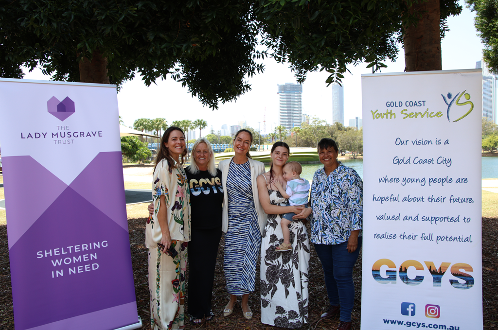 Four women, one holding a baby posing for a photo between the LMT and GCYS banners
